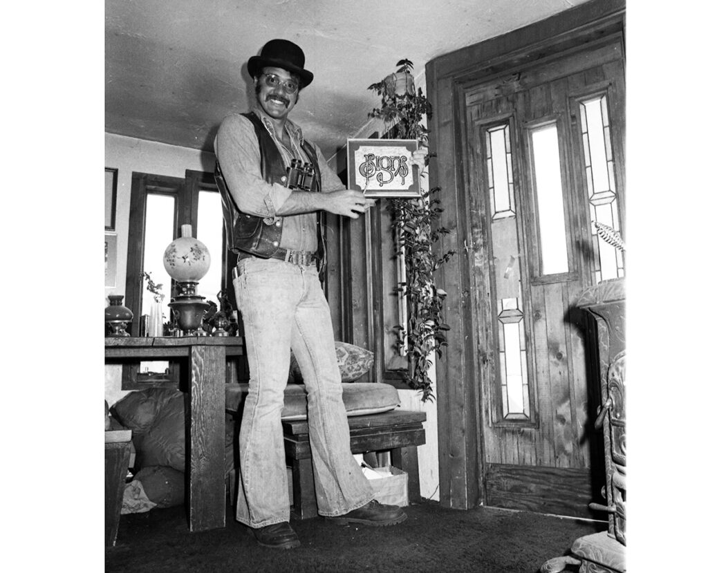 One 35 mm b/w film negative of Gaard Moses, 1979. He is holding a sign that he made, and it reads "Signs" in decorative letters. He has a pair of binoculars around his neck and is wearing bell-bottom jeans.