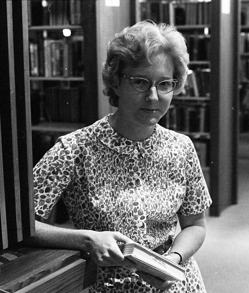 Image of a woman standing next to book shelves holding a book in her hands. She is wearing a button down printed dress and cat eye glasses with short feathered hair.