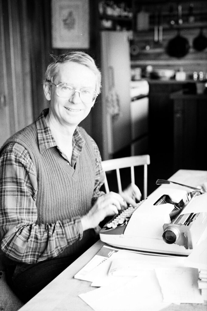 Image of a man wearing a button up shirt under a sweater vest sitting at a table with his hands resting on a typewriter with a kitchen in the background.